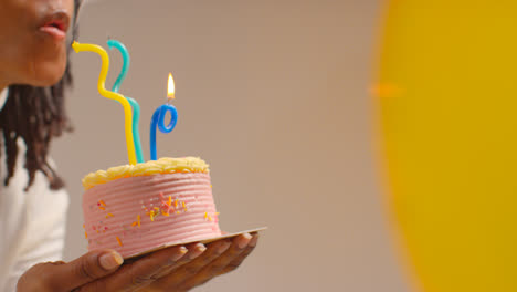 Close-Up-Studio-Shot-Of-Woman-Wearing-Birthday-Headband-Celebrating-Birthday-Blowing-Out-Candles-On-Cake